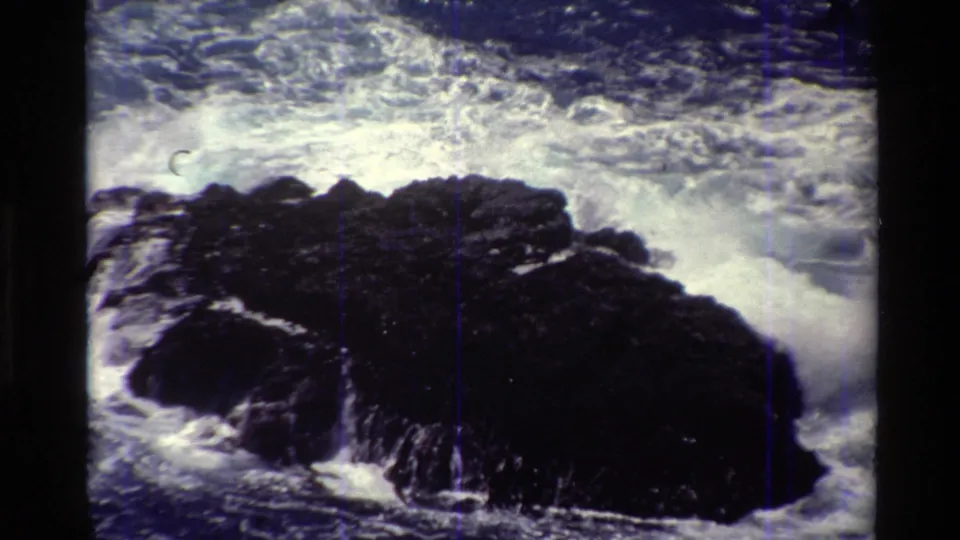 Big Sur coastal frame with dark rocks surrounded by turbulent white foam, recorded on grainy 8mm film.