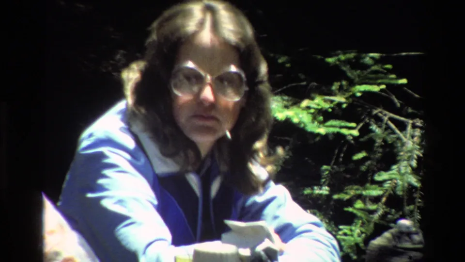 In 1980 Big Sur footage, a woman wearing large glasses and a blue jacket sits at an outdoor picnic spot near evergreen foliage.