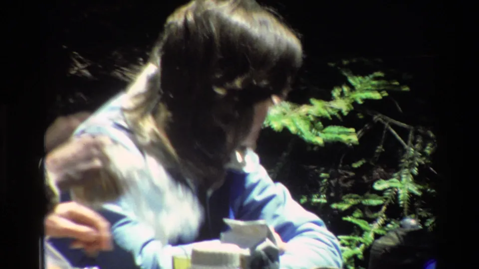 The woman in the blue jacket turns aside at the Big Sur picnic spot, with small items and evergreen branches visible around her.