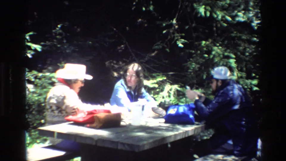 Three campers at a wooden picnic table in Big Sur, California, with bags and cups visible in bright forest light.
