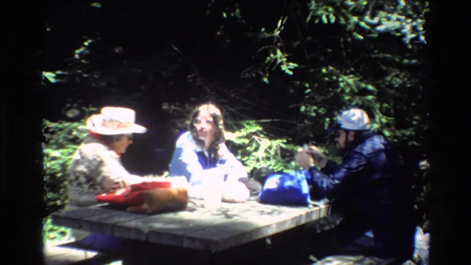 Three campers remain at a Big Sur picnic table, partially overexposed by sun and surrounded by dark forest shade.