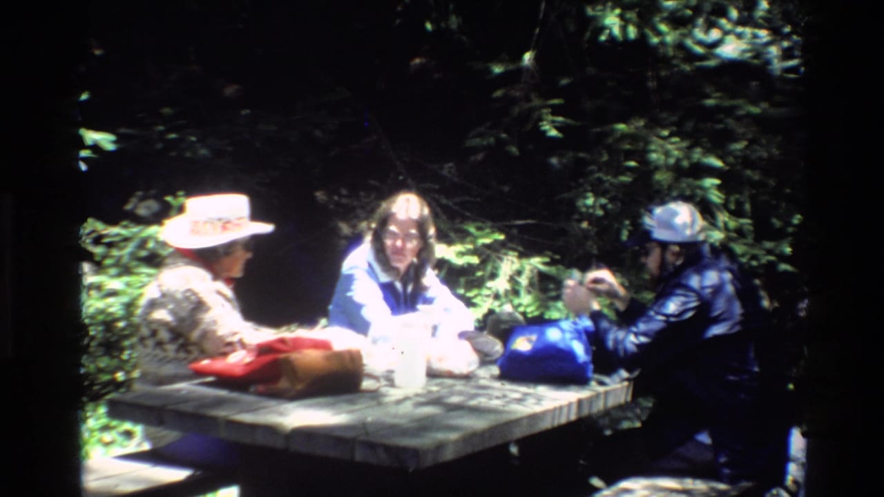1980 Big Sur home-movie frame showing three adults at a wooden picnic table under trees, with hats, jackets, and dappled sunlight.