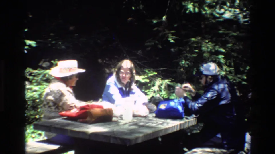 1980 Big Sur home-movie frame showing three adults at a wooden picnic table under trees, with hats, jackets, and dappled sunlight.