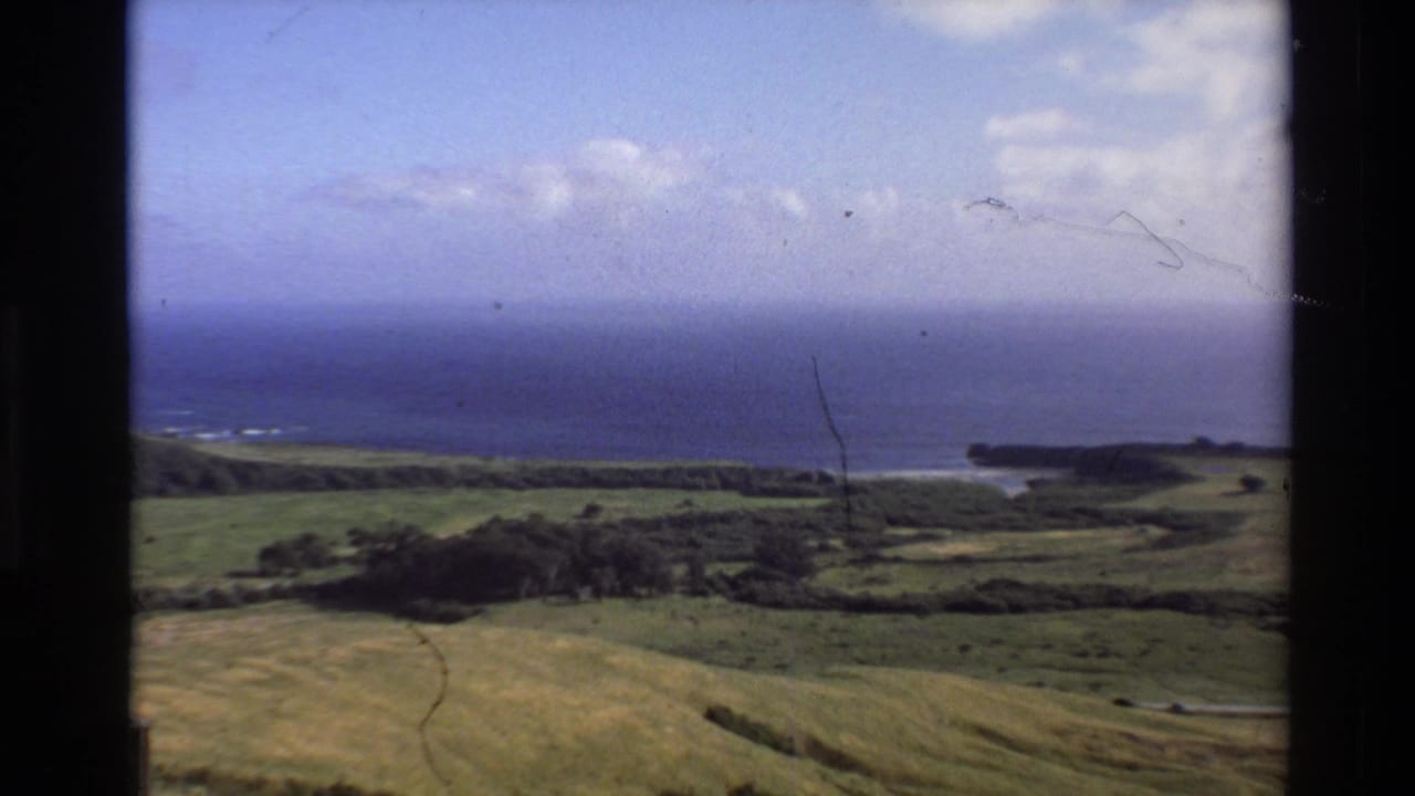 1980 Big Sur coastal landscape frame with green hills, Pacific Ocean, distant shoreline, blue sky, and visible film wear.