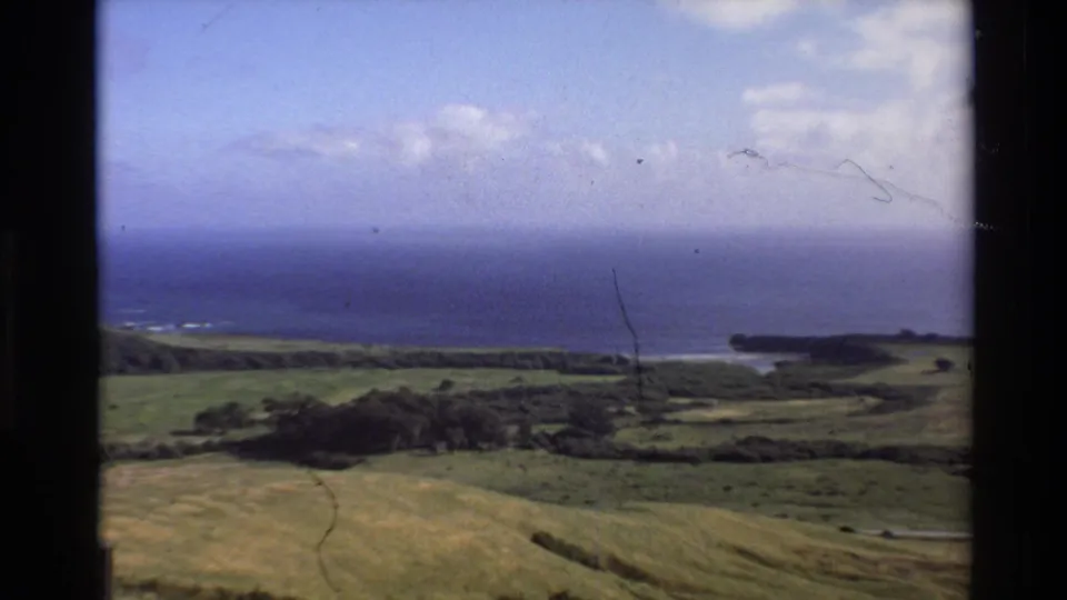 1980 Big Sur coastal landscape frame with green hills, Pacific Ocean, distant shoreline, blue sky, and visible film wear.