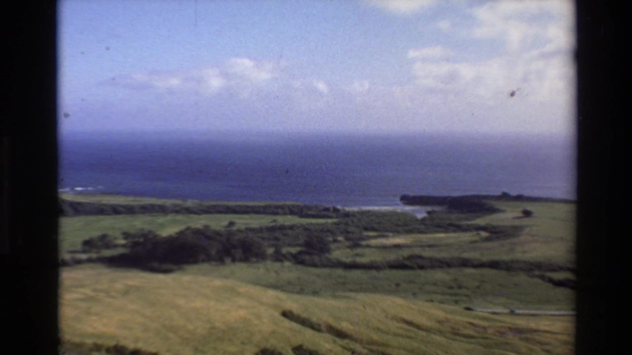 1980 8mm view of Big Sur green countryside leading toward blue Pacific water and a distant bluff under scattered clouds.