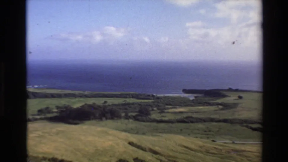 1980 8mm view of Big Sur green countryside leading toward blue Pacific water and a distant bluff under scattered clouds.