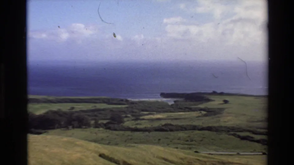 Big Sur coastal countryside in 1980 8mm footage, with green fields, ocean horizon, and dark bluffs framed by film gate edges.