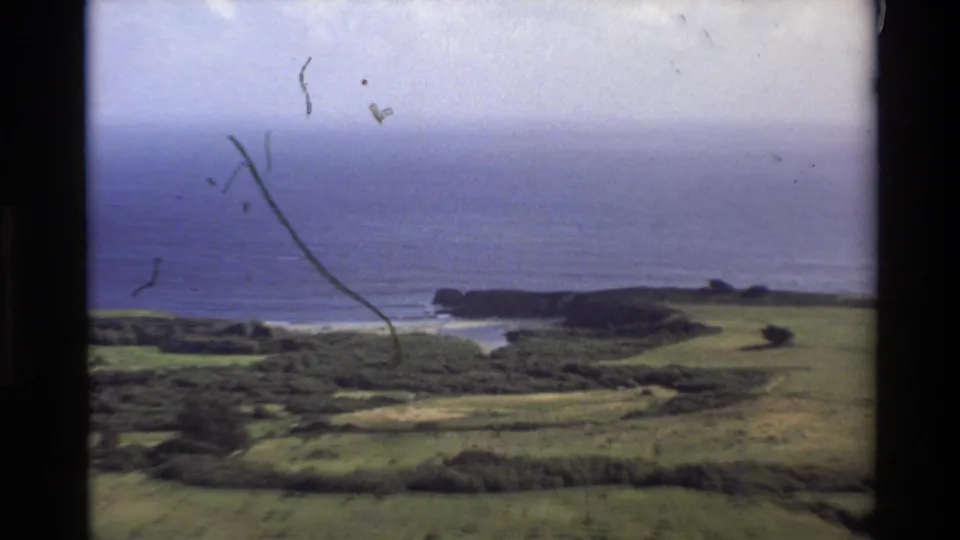 Closer 1980 8mm view of the Big Sur shoreline, showing green fields, wooded patches, blue water, and pale surf near a rocky bluff.