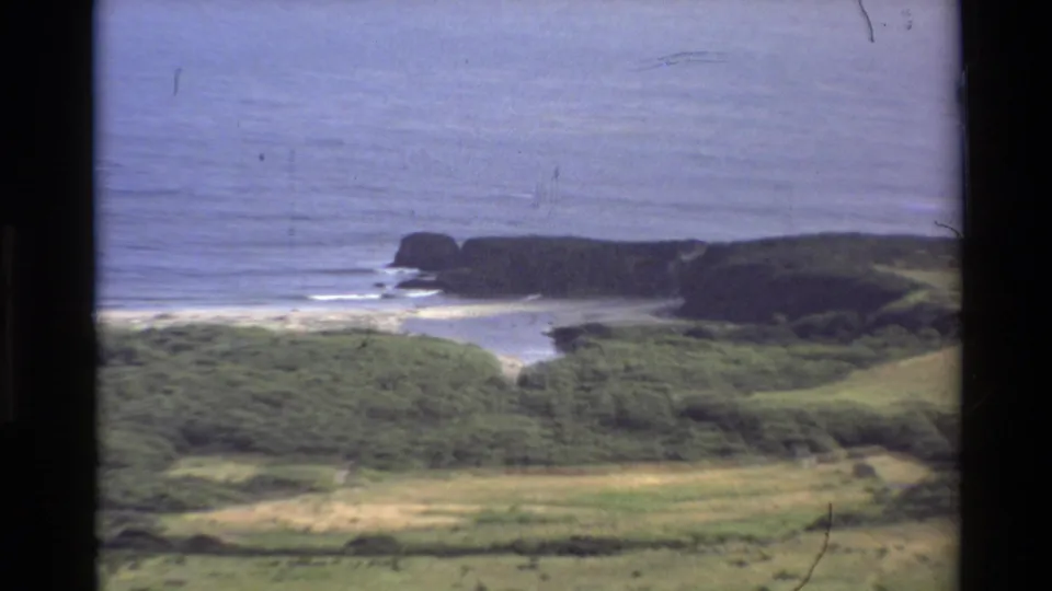 1980 8mm Big Sur shoreline detail with a rocky alcove, pale surf, green foreground slopes, and calm blue Pacific water.