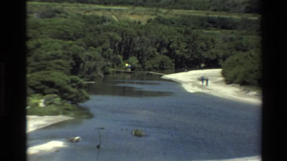 Big Sur-area home-movie frame showing a calm river or estuary, dense green vegetation, and distant people on a light sandbank.