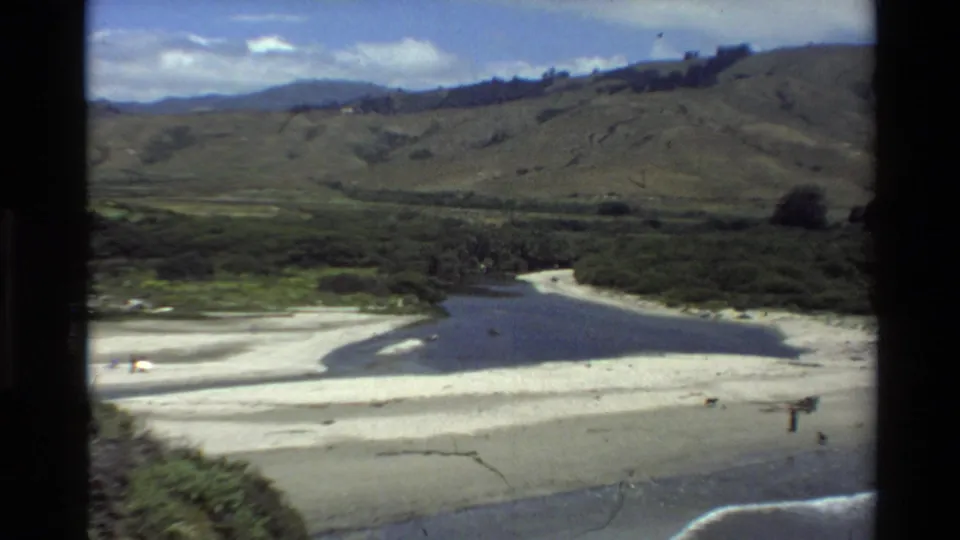 Big Sur, California aerial frame showing pale sand, a dark river mouth, low surf, hills, and tiny beach visitors in 1980 footage.