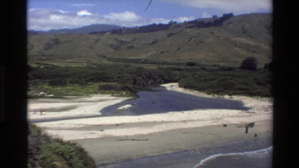 1980 Big Sur aerial frame with a river channel crossing pale beach sand toward ocean surf beneath broad mountain slopes.