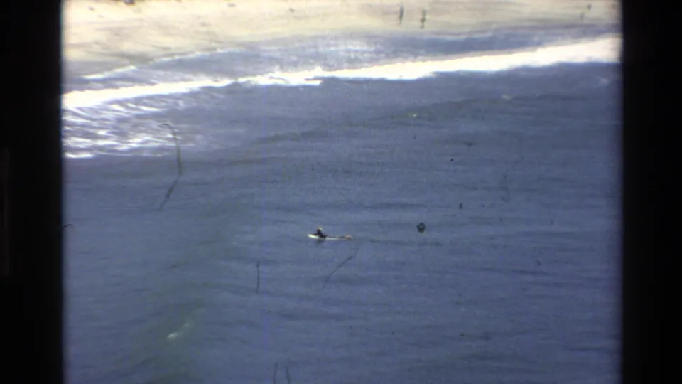 A distant surfer paddles in blue ocean water below a line of white surf along the Big Sur coast.