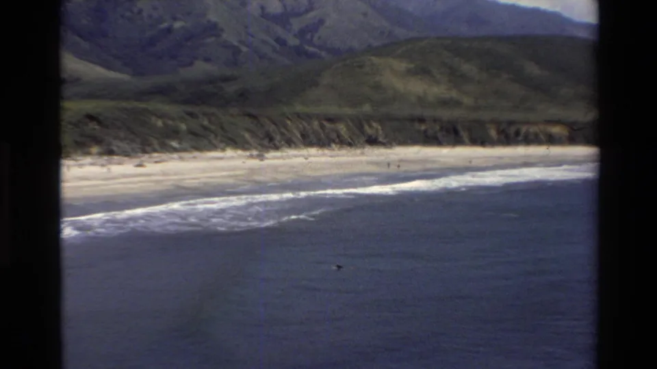 Wide 1980s 8mm view of Big Sur coastline, showing pale sand, breaking waves, green hills, and a small surfer offshore.