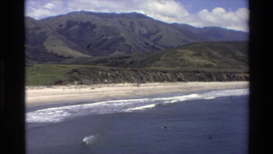 Big Sur coastline in 1980s 8mm footage, with a pale beach, rolling surf, green mountains, and a small surfer offshore.