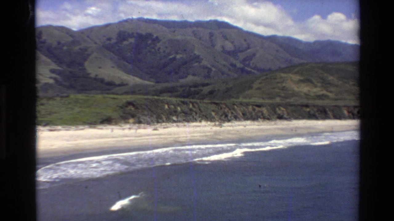 Waves roll onto a sandy beach below green coastal mountains.