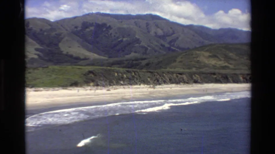 1980 Big Sur 8mm frame showing surf on a sandy beach with green mountains and cloudy sky behind it.