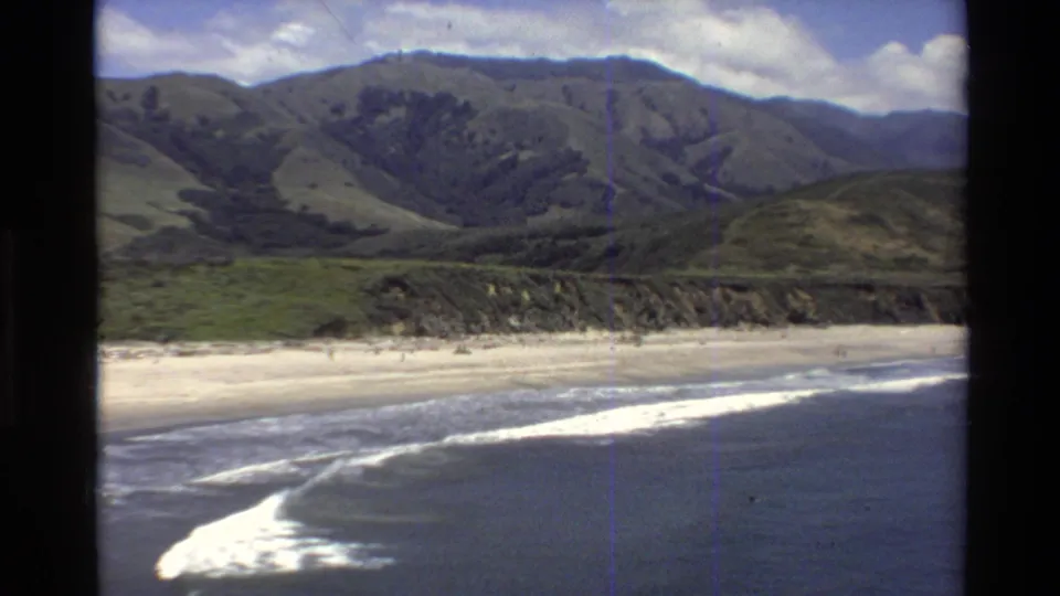 8mm home-movie frame of the Big Sur coast, with a sandy beach, surf line, distant people, and green mountain slopes.