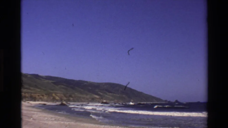 Big Sur shoreline frame showing beach-level surf, distant coastal hills, open blue sky, and visible film wear.