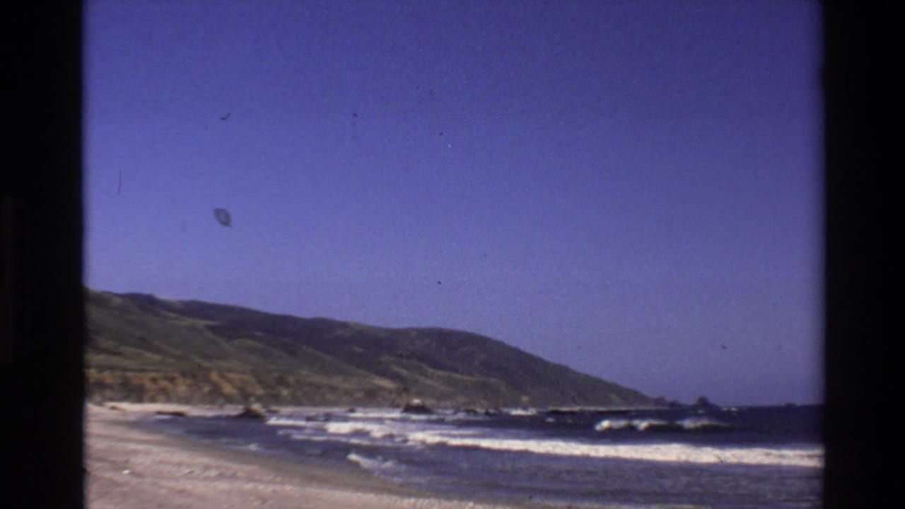Waves rolling onto a sandy beach below a coastal hillside and blue sky.