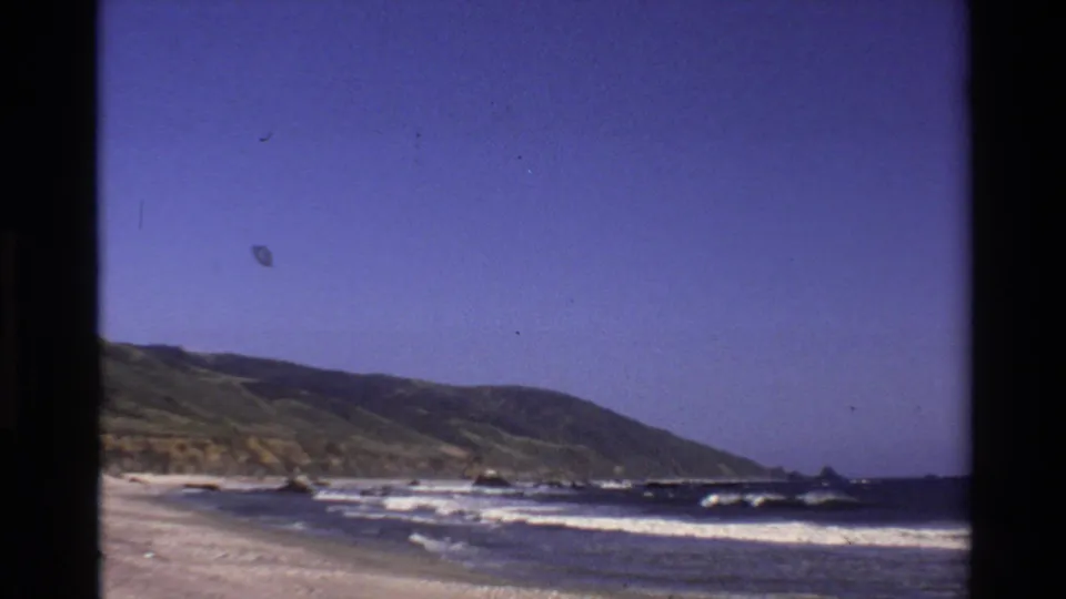 1980 8mm home-movie frame of Big Sur coastline with surf, sand, offshore rocks, hillside, and blue sky.