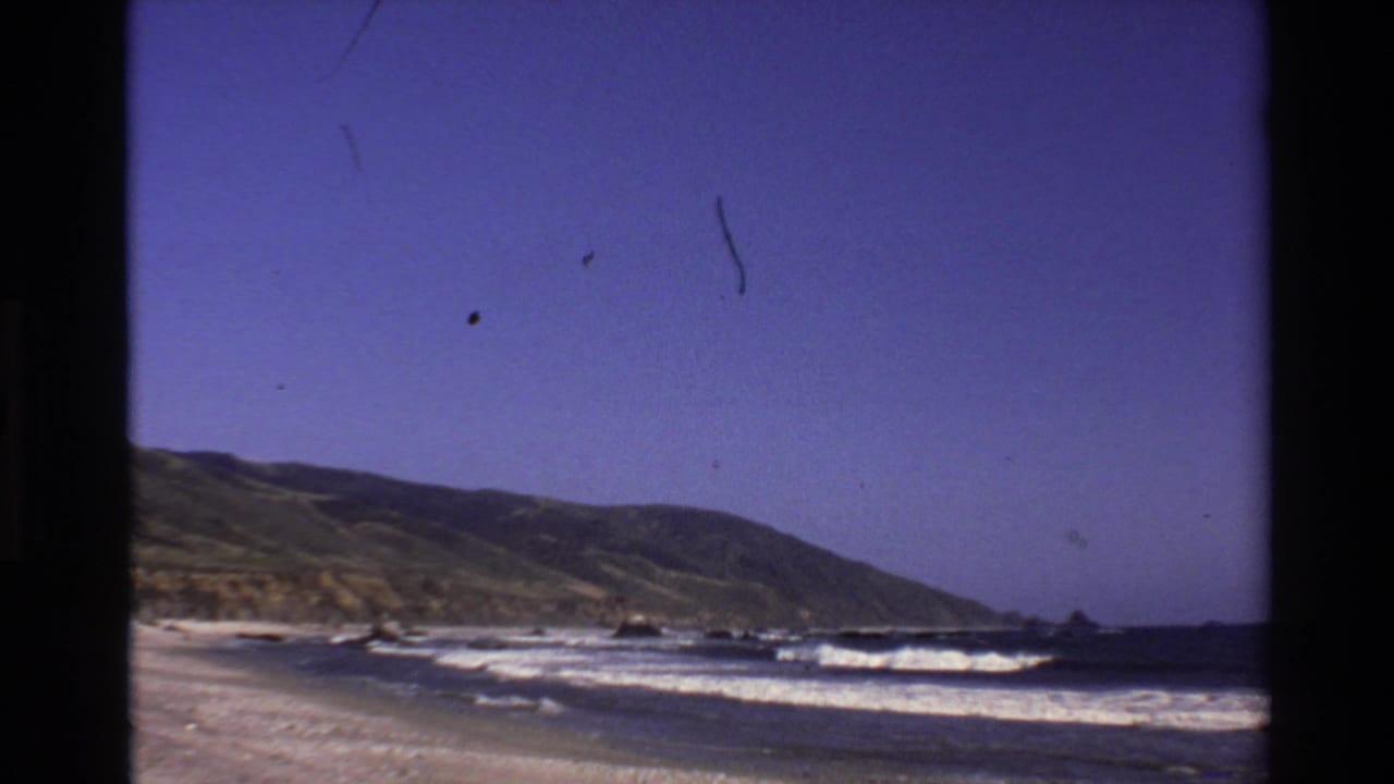 1980 home-movie view of the Big Sur shore, with surf lines, beach sand, scattered rocks, and a distant hillside.