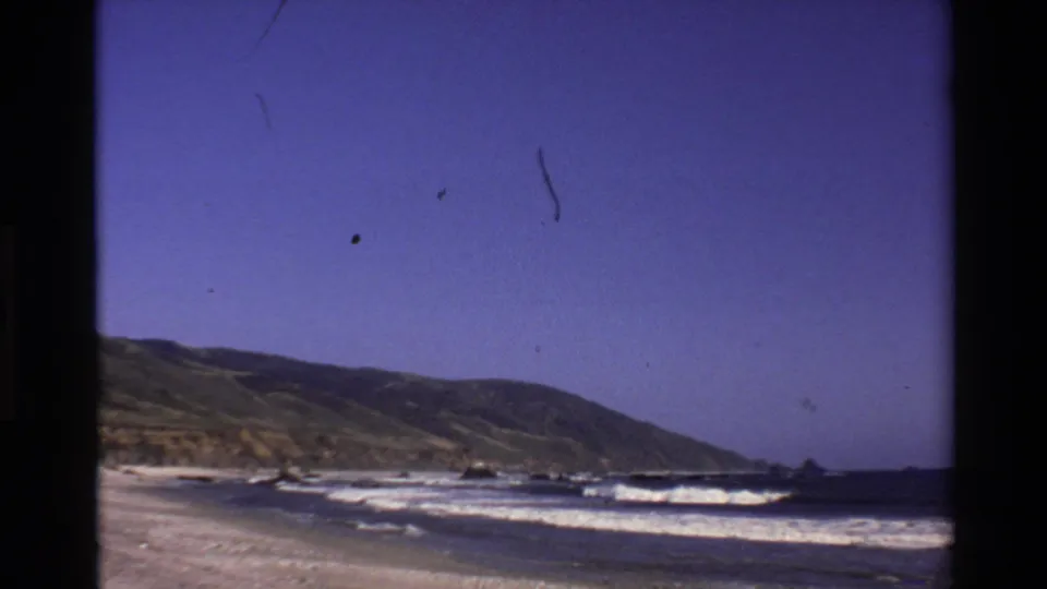 1980 home-movie view of the Big Sur shore, with surf lines, beach sand, scattered rocks, and a distant hillside.
