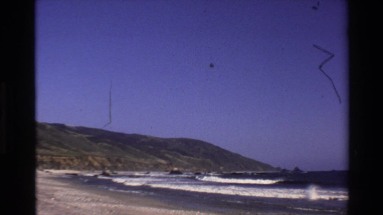 Big Sur coastal home-movie frame from 1980 showing white surf, sandy beach, dark rocks, hillside, and visible film wear.