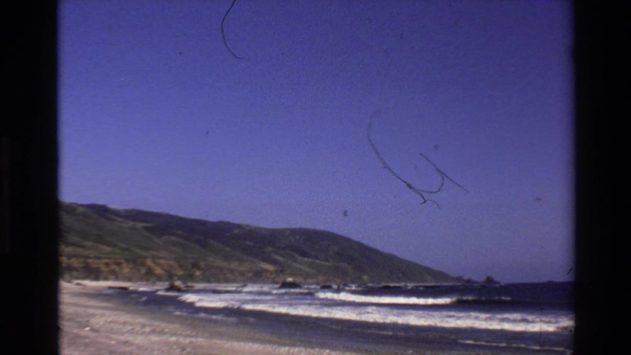 1980 Big Sur 8mm frame showing surf breaking along a sandy beach with a green hillside and blue sky beyond.