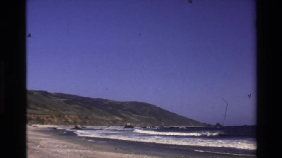 Big Sur coastal view from 1980 8mm footage, with white waves crossing the shore below a green hillside.