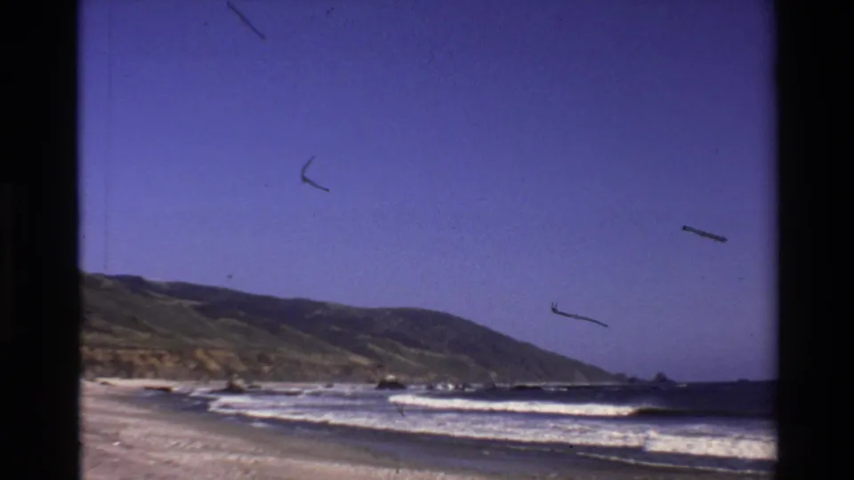 1980 Big Sur home-movie frame showing beach, rolling waves, and a distant green coastal slope beneath blue sky.