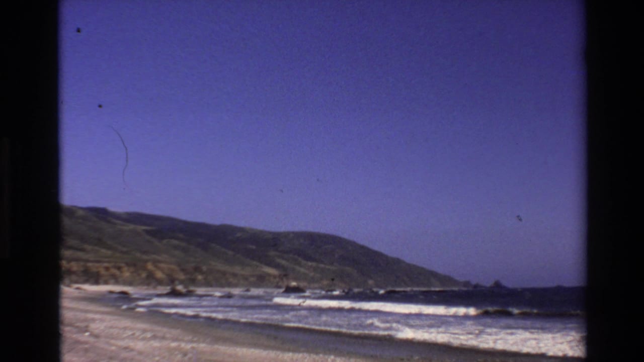 Wide coastal beach view with waves, green hillside, blue sky, and dark film borders.