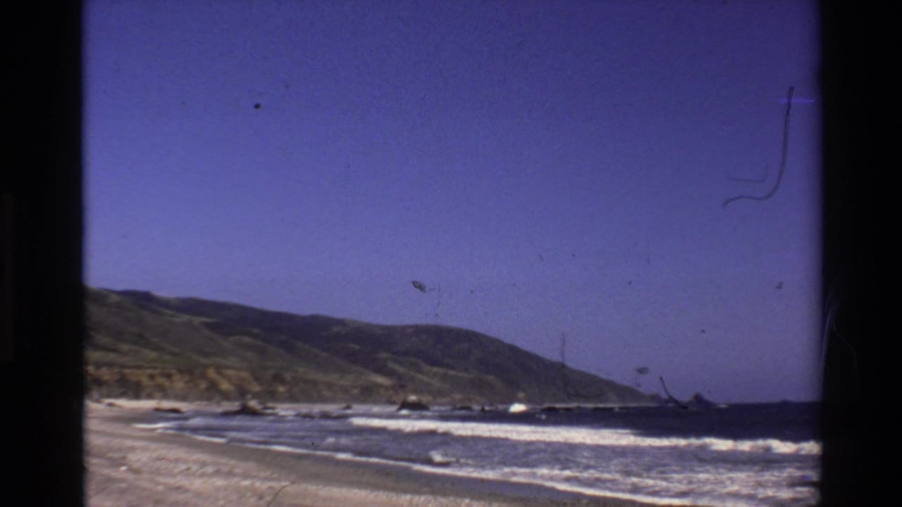 Vintage 8mm view of the Big Sur coast with waves reaching a sandy beach beneath low green hills.