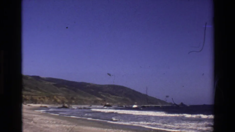 Vintage 8mm view of the Big Sur coast with waves reaching a sandy beach beneath low green hills.