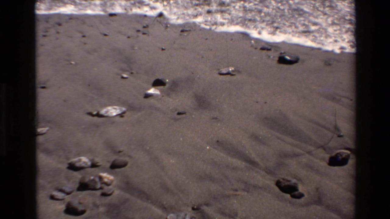 Closer 1980 Big Sur beach frame with pebbles on wet sand, foamy surf, ocean, and coastal hillside in the background.