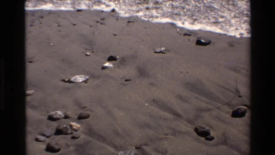 Closer 1980 Big Sur beach frame with pebbles on wet sand, foamy surf, ocean, and coastal hillside in the background.