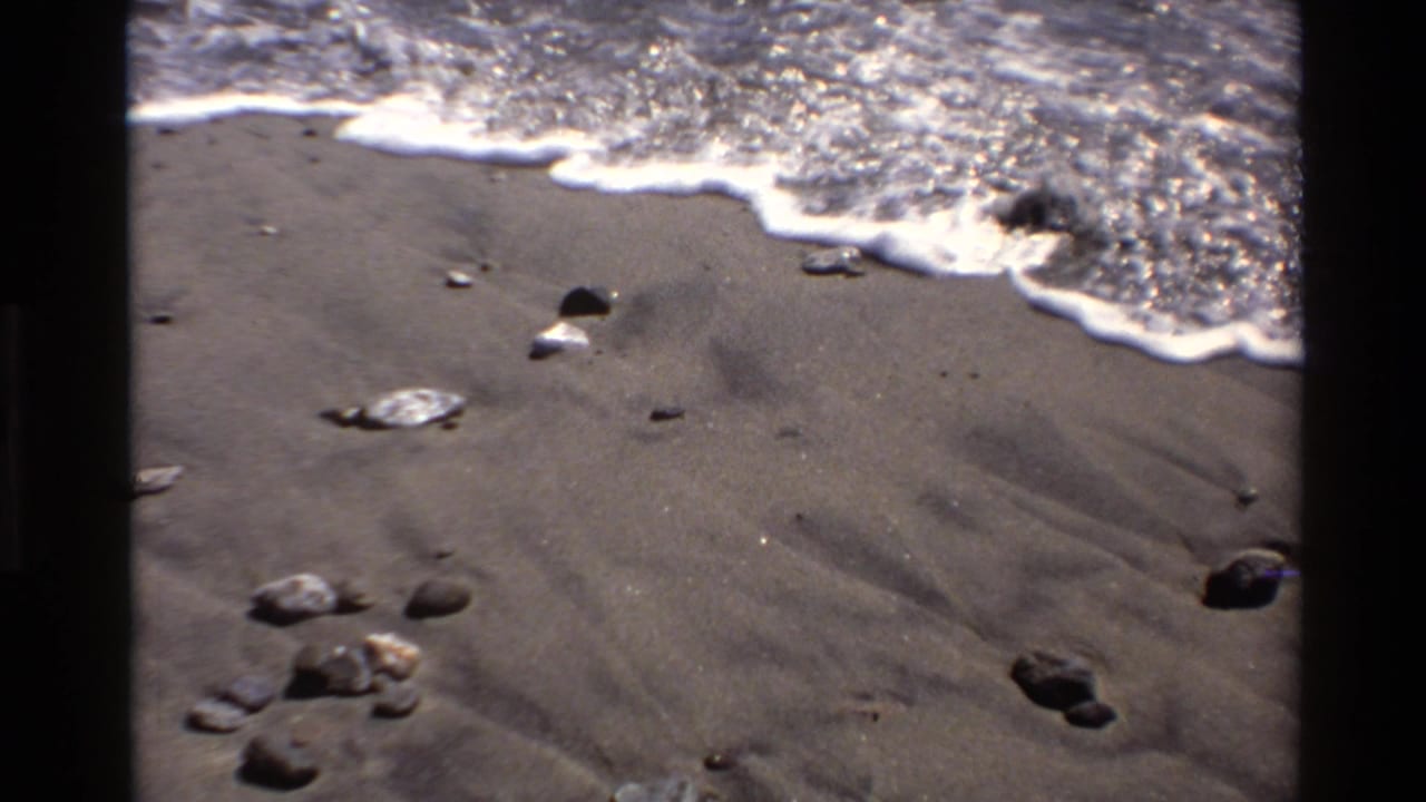 1980 Big Sur 8mm frame showing white surf edging across dark beach sand and small rocks in bright sun.