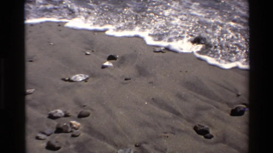 1980 Big Sur 8mm frame showing white surf edging across dark beach sand and small rocks in bright sun.
