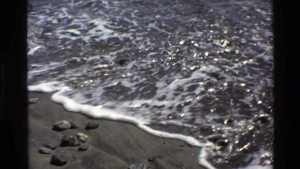 1980 Big Sur shoreline frame with rippled seawater and foam washing over dark sand beside scattered rocks.