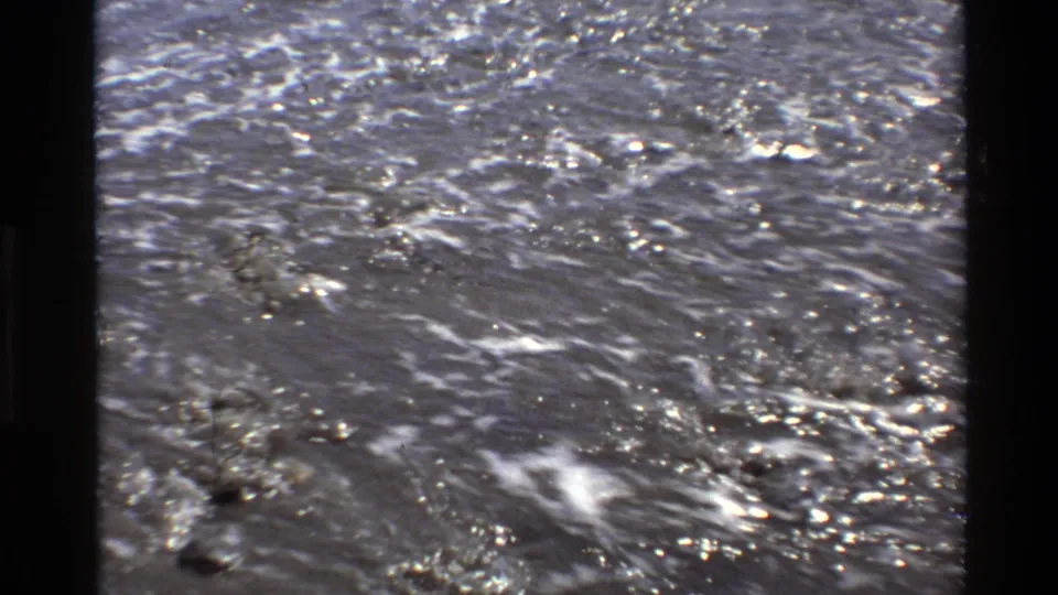 1980 Big Sur coastal frame showing shimmering seawater and sparse foam after a wave has covered the sand.