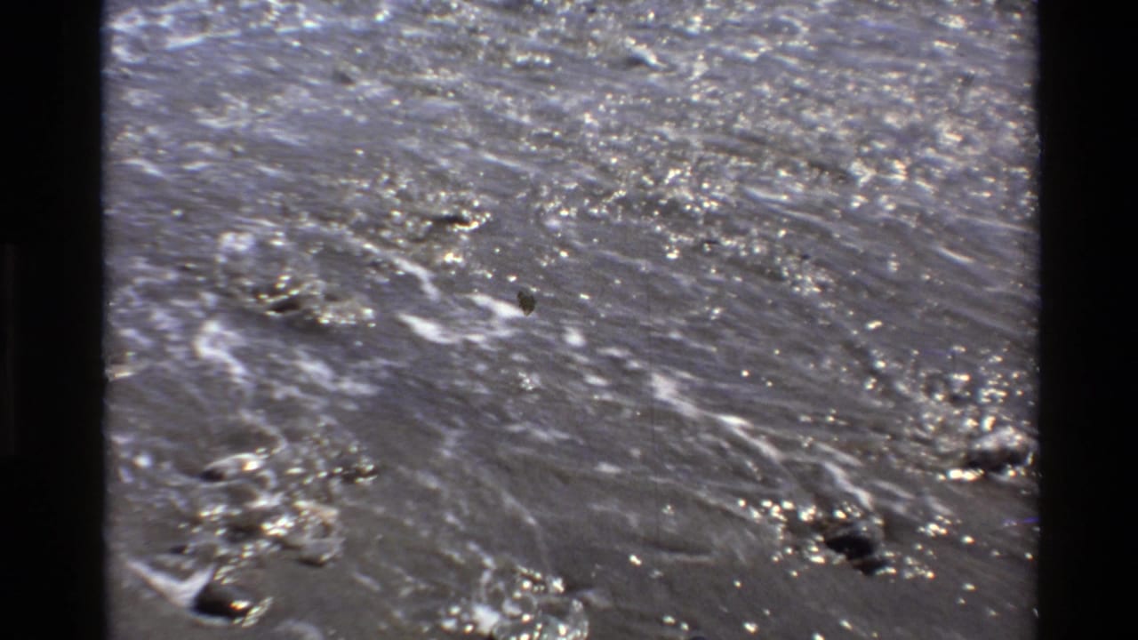 1980 Big Sur 8mm frame showing close water movement across wet shoreline sand, scattered stones, bubbles, and glinting highlights.