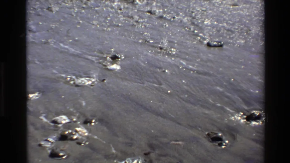 1980 Big Sur shoreline detail with thin water flow across dark sand, scattered stones, and visible 8mm frame border.