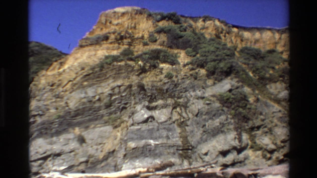 1980 Big Sur coastal cliff frame showing layered rock faces, scrub vegetation, and blue sky in a handheld 8mm home-movie view.
