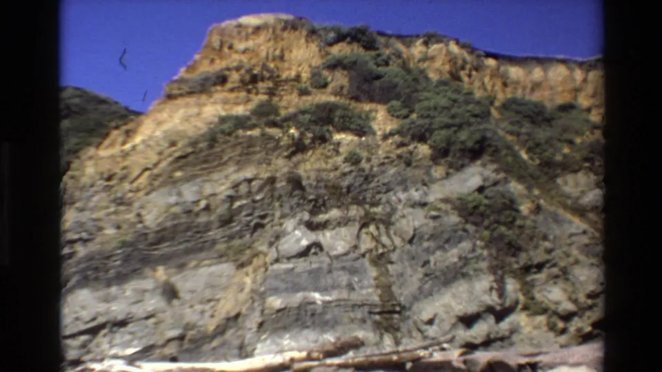 1980 Big Sur coastal cliff frame showing layered rock faces, scrub vegetation, and blue sky in a handheld 8mm home-movie view.