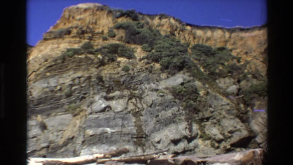 1980 Big Sur frame with a rugged layered bluff, scattered shrub growth, blue sky, and visible 8mm frame edges.