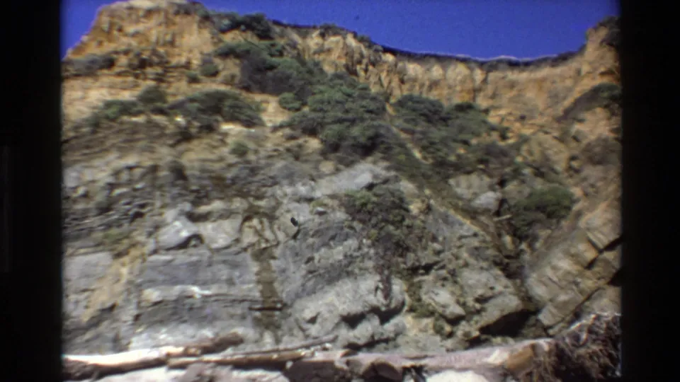 1980 Big Sur home-movie view of a rocky cliff with scrub vegetation, pale shore edge, and driftwood at the base.