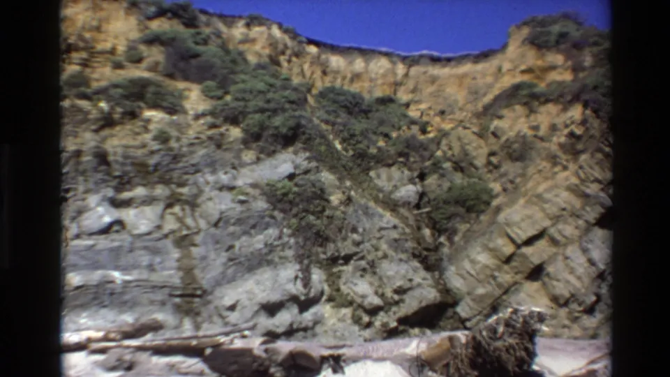 1980 Big Sur frame showing a rugged stratified cliff, shrub-covered slopes, blue sky, and driftwood near the lower edge.