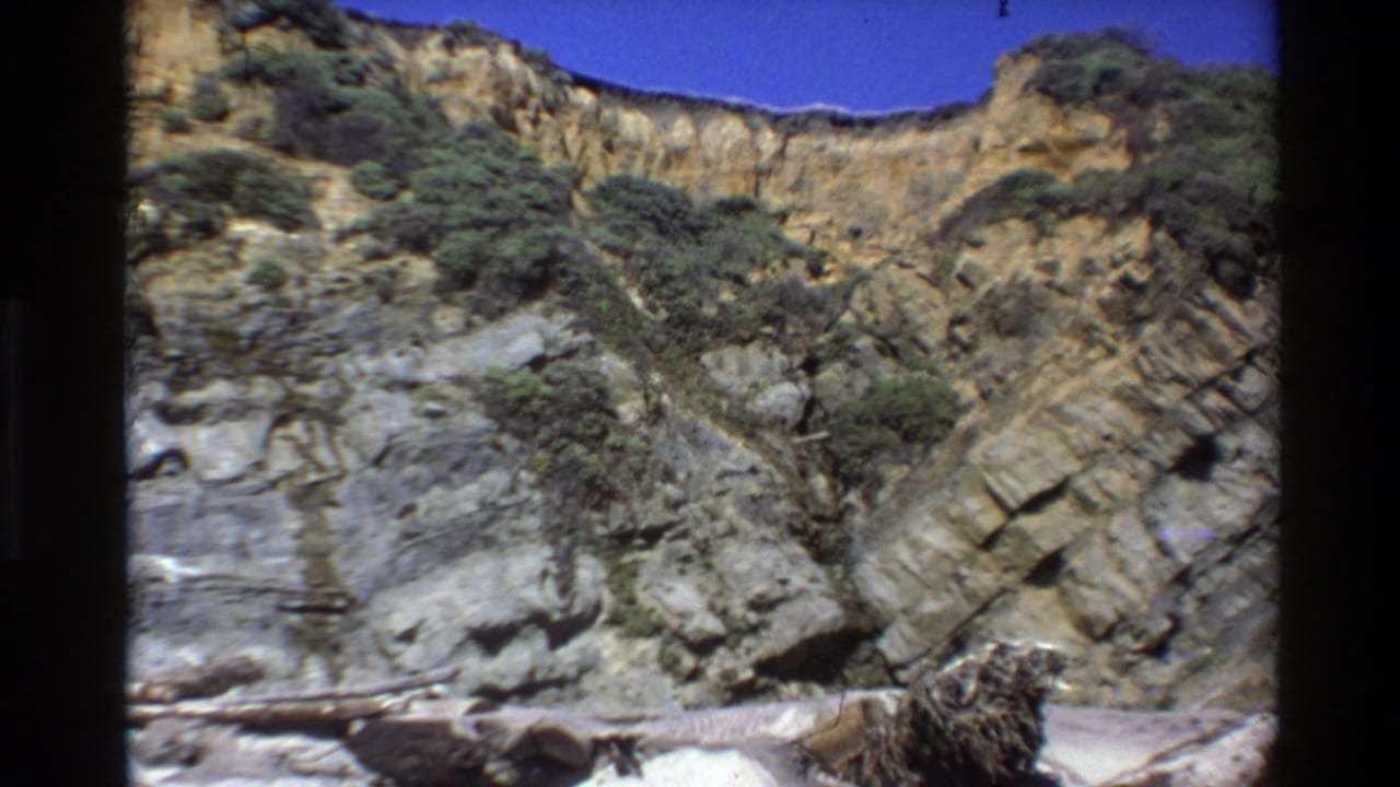 Big Sur, California 8mm frame showing a layered rocky cliff, scattered green shrubs, blue sky, and pale shoreline debris.