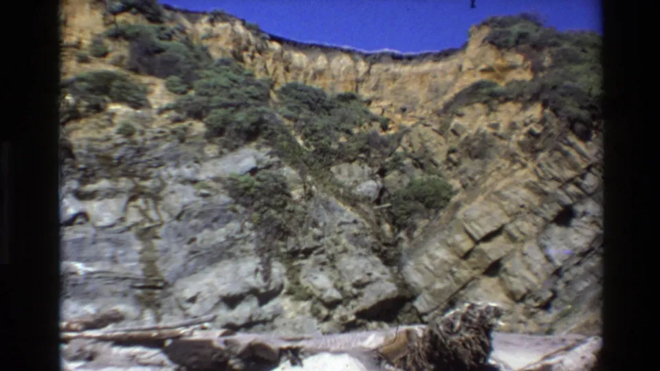 Big Sur, California 8mm frame showing a layered rocky cliff, scattered green shrubs, blue sky, and pale shoreline debris.